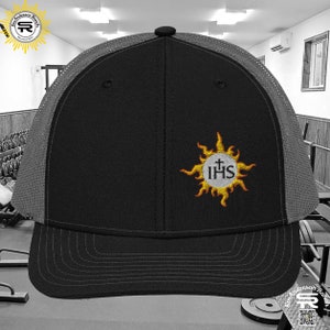 Puede incluir: Gorra de camionero negra y gris con una parte trasera de malla negra. La parte delantera de la gorra presenta un sol amarillo bordado con un círculo blanco que contiene una cruz y las letras "IHS".