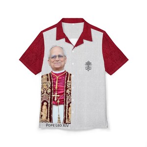 Può includere: Una camicia a maniche corte con un ritratto di Papa Leone XIV. La camicia ha un corpo bianco con un motivo sottile e maniche e colletto bordeaux. Il testo "POPE LEO XIV" è stampato in basso.
