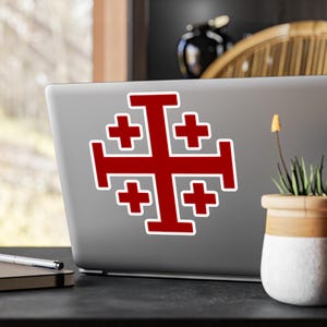 Pode incluir: Um adesivo de cruz de Jerusalém vermelho em um laptop prateado. A cruz é um símbolo do cristianismo e é frequentemente usada na arte e na arquitetura religiosas.