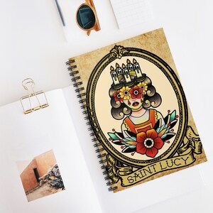 May include: A vintage-style illustration of Saint Lucy, a female figure with candles on her head and flowers around her eyes. The image is framed in a gold oval with the text "Saint Lucy" at the bottom.