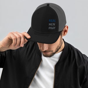 Puede incluir: Una gorra de camionero negra y gris con una visera negra y una malla gris en la parte posterior. La gorra tiene las palabras "REAL MEN PRAY" bordadas en hilo azul y blanco.