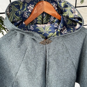 May include: A hooded cape made of a blue and white herringbone tweed fabric. The hood is lined with a floral patterned fabric in shades of green, white, and blue. A decorative metal clasp is attached to the front of the cape.