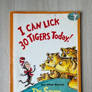 May include: A Dr. Seuss paperback classic book cover featuring a colorful illustration of the Cat in the Hat and a group of tigers. The title of the book is "I Can Lick 30 Tigers Today!" and the author is Dr. Seuss.