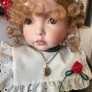May include: A porcelain doll with curly blonde hair and brown eyes. The doll wears a green dress with red apple patterns, a white collar with lace trim, and a gold locket necklace. A red rose adorns the dress.