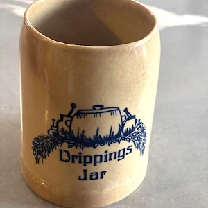 May include: A tan ceramic jar with a blue illustration and the words "Drippings Jar" in blue. The jar has a cylindrical shape and a wide opening. The illustration depicts a pot and other kitchen items.