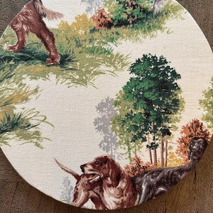 May include: A round placemat with a hunting scene featuring two brown and black dogs, trees, and green grass on a cream background.