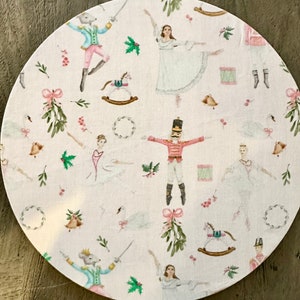May include: A round white tablecloth with a festive Nutcracker theme. The tablecloth features a repeating pattern of ballerinas, nutcrackers, toy soldiers, rocking horses, and mistletoe. The design is in shades of pink, green, and white.