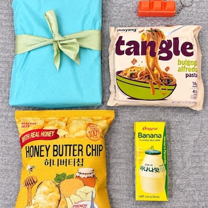 May include: A light blue gift wrapped in paper with a light green ribbon, an orange Tetris-style keychain, a package of Samyang Tangle bulgogi alfredo pasta, a bag of Haitai Honey Butter Chips, and a carton of Binggrae Banana flavored milk drink.