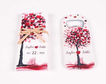 May include: A set of wedding favors featuring a white background with a red and pink tree design. The tree is made up of hearts and has the names "Angelica" and "Austin" written on it. The date "03 | 22 | 2024" is also written on the favor. One favor is a small box tied with a brown ribbon and the other is a bottle opener.
