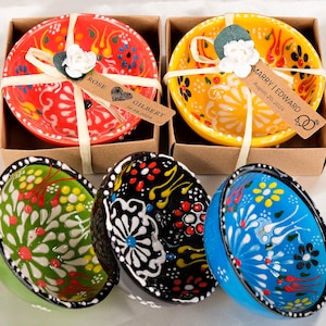 May include: A selection of brightly coloured ceramic bowls featuring floral designs. Two bowls are presented in brown boxes, tied with ribbon, and labelled with tags. The tags read "Rose Gilbert 10.08.2024" and "Marry Edward August 20, 2024". The bowls are red, yellow, green, black, and blue.