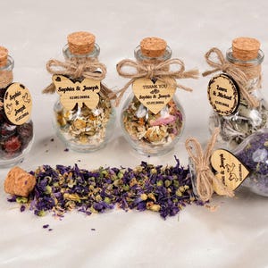 May include: A collection of small glass bottles filled with dried herbs and flowers, sealed with corks and twine. Wooden tags with names and dates are attached. Some tags are butterfly or heart-shaped. The text on the tags reads "Sophia & Joseph" and "Laura & Michael".