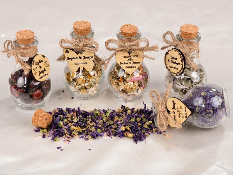 May include: A collection of small glass bottles filled with dried herbs and flowers, sealed with corks and twine. Wooden tags with names and dates are attached. Some tags are butterfly or heart-shaped. The text on the tags reads "Sophia & Joseph" and "Laura & Michael".