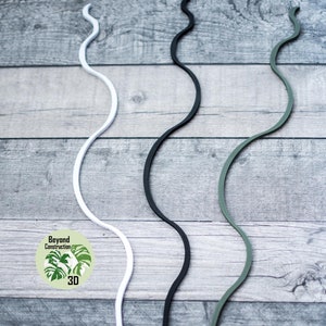 May include: Three plant supports made of flexible plastic. The supports are white, black, and green. They are all in a wavy shape and are lying on a wooden surface.