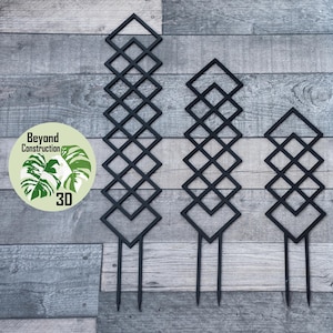 May include: Three black metal plant stakes with a diamond pattern. The stakes are different sizes and are arranged in a row on a wood background.