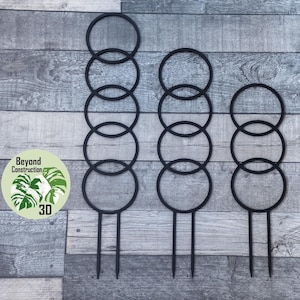 May include: Three black metal plant supports with circular rings. The supports are different heights, with the tallest having five rings, the middle having four rings, and the shortest having three rings.