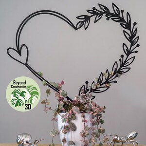May include: A black metal heart-shaped wreath with leafy details sits behind a potted plant with green and pink leaves. A silver snail and ant figurine are on a wooden surface below the plant.