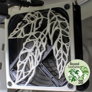 May include: A white 3D printed fan grill with a leaf design. The grill is attached to a black computer fan. The text "Beyond Construction 3D" is printed on a green sticker.