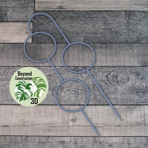 May include: A grey 3D-printed plant support with three circular openings and pointed ends, on a wooden surface. A circular sticker with the text "Beyond Construction 3D" and a plant graphic is visible.