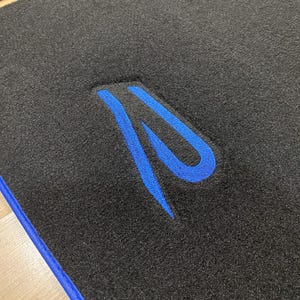 May include: Black car floor mat with a blue embroidered design. The mat has a blue border and a textured surface. The design is a stylized letter "R".