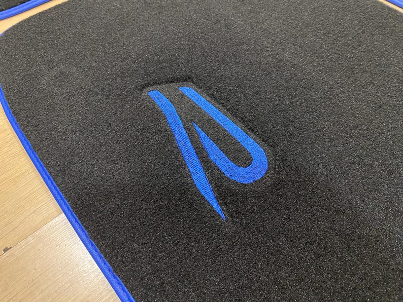 May include: Black car floor mat with a blue embroidered design. The mat has a blue border and a textured surface. The design is a stylized letter "R".