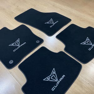 May include: Set of four black car floor mats with the white "CUPRA" logo embroidered on each mat. The mats are shaped to fit the car's floor and have a non-slip backing.