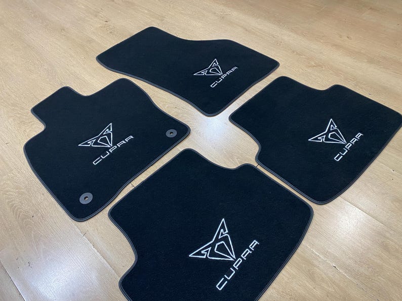 May include: Set of four black car floor mats with the white "CUPRA" logo embroidered on each mat. The mats are shaped to fit the car's floor and have a non-slip backing.