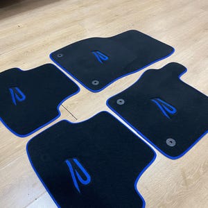 May include: Set of four black car floor mats with blue trim and a blue embroidered design. The mats are designed to fit a vehicle's front and rear footwells. The mats have a rubber backing and are designed to protect the vehicle's carpet.