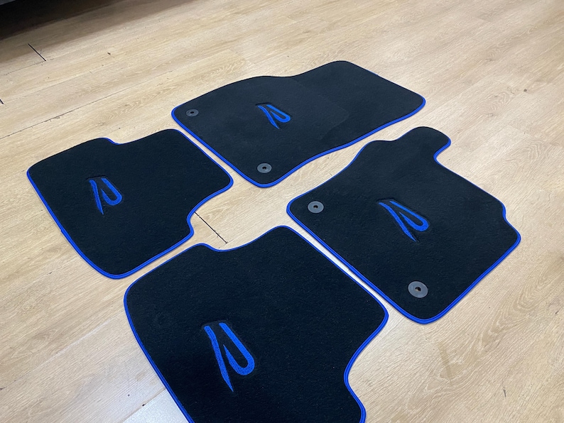 May include: Set of four black car floor mats with blue trim and a blue embroidered design. The mats are designed to fit a vehicle's front and rear footwells. The mats have a rubber backing and are designed to protect the vehicle's carpet.