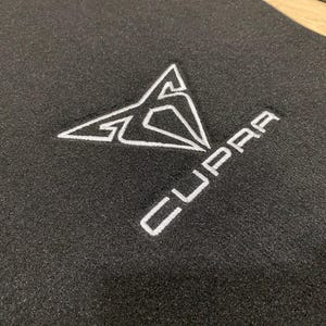 May include: Black car floor mat with a white embroidered Cupra logo. The logo is a stylized geometric design with the brand name below it. The mat is made of a textured material.