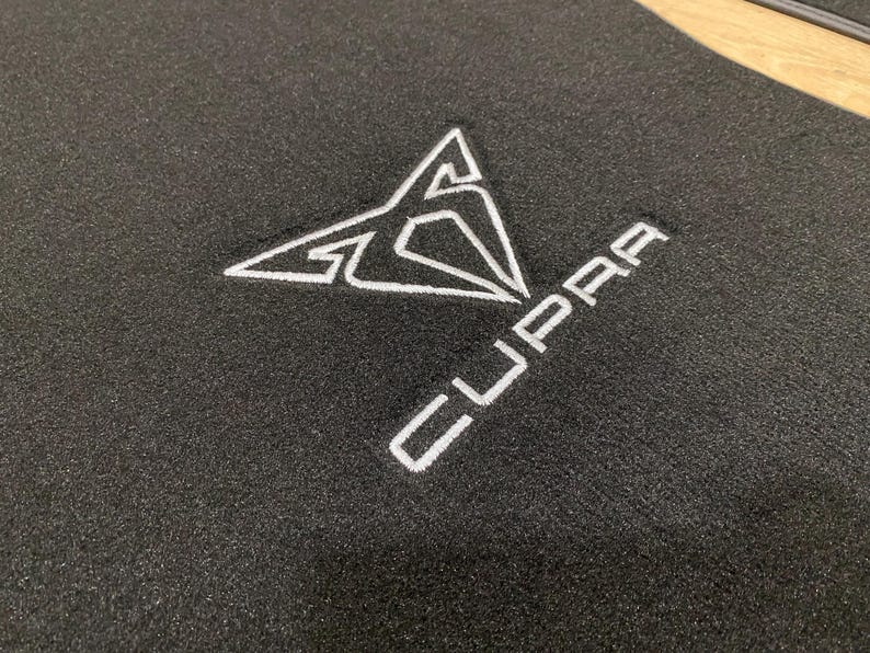 May include: Black car floor mat with a white embroidered Cupra logo. The logo is a stylized geometric design with the brand name below it. The mat is made of a textured material.