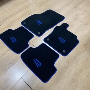 May include: Set of four black car floor mats with blue trim and a stylized blue letter "R" embroidered on each mat. The mats are arranged on a light wood surface.