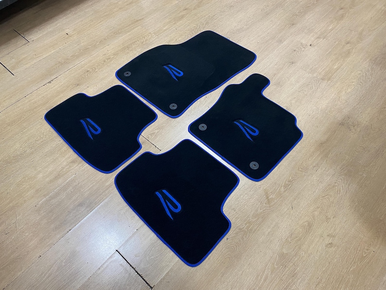 May include: Set of four black car floor mats with blue trim and a stylized blue letter "R" embroidered on each mat. The mats are arranged on a light wood surface.