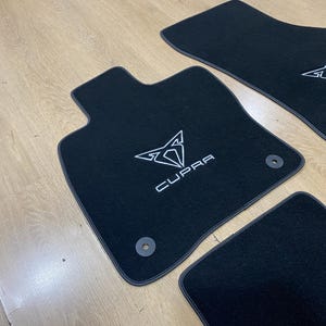May include: Black car floor mats with the white "CUPRA" logo and emblem. The mats have a dark gray trim and are arranged on a light wood surface. The mats are designed to protect the car's interior.