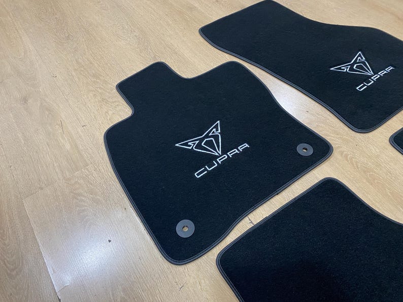 May include: Black car floor mats with the white "CUPRA" logo and emblem. The mats have a dark gray trim and are arranged on a light wood surface. The mats are designed to protect the car's interior.