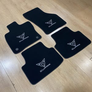 May include: Set of four black car floor mats with a white Cupra logo. The mats are arranged on a light wood surface. The mats are shaped to fit the car's footwells.