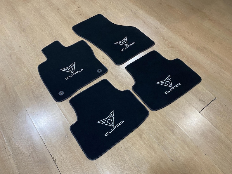 May include: Set of four black car floor mats with a white Cupra logo. The mats are arranged on a light wood surface. The mats are shaped to fit the car's footwells.