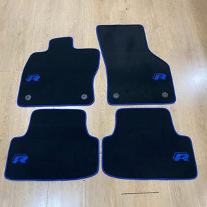 Könnte beinhalten: Vier schwarze Auto-Fußmatten mit blauer Umrandung. Jede Matte hat ein blaues "R"-Logo. Die Matten sind für die vorderen und hinteren Fußräume eines Fahrzeugs konzipiert. Hergestellt aus strapazierfähigem Material.