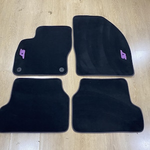 May include: A set of four black automotive floor mats with a soft, carpeted texture. Two mats feature a purple embroidered ST logo. The front mats include circular mounting grommets, while the rear mats have a rectangular shape with rounded edges.