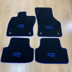Könnte beinhalten: Vier schwarze Auto-Fußmatten mit blauer Umrandung und blauem "R"-Logo. Die Matten sind für den Fahrzeuginnenraum konzipiert und verfügen über Befestigungen.