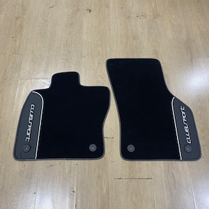 Puede incluir: Dos alfombrillas de coche negras con una forma asimétrica distintiva. Cada alfombrilla presenta un panel de acento gris a lo largo del borde exterior con el texto "CLUBSPORT" impreso en blanco. Incluyen ojales circulares para su instalación.
