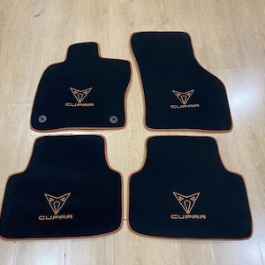 May include: Set of four black car floor mats. Each mat features the Cupra logo in a copper color and the word "CUPRA" below the logo. The mats have a copper-colored border and are designed to fit a car.