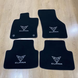 May include: Set of four black car floor mats. Each mat features the white "CUPRA" logo and emblem. The mats are designed to fit a vehicle's interior and protect the floor.