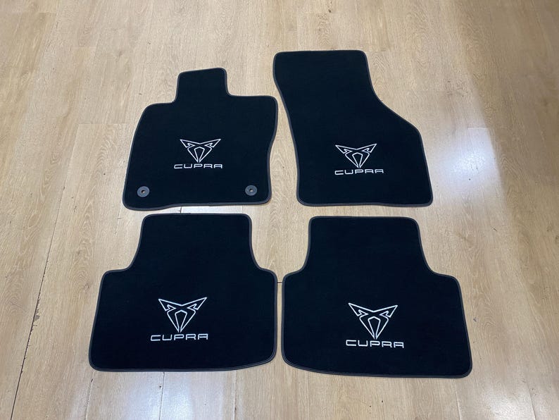 May include: Set of four black car floor mats. Each mat features the white "CUPRA" logo and emblem. The mats are designed to fit a vehicle's interior and protect the floor.