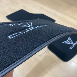 May include: Black car floor mats with the white "CUPRA" logo embroidered on the surface. The mats have a dark gray trim and are designed to protect the vehicle's interior from dirt and wear.
