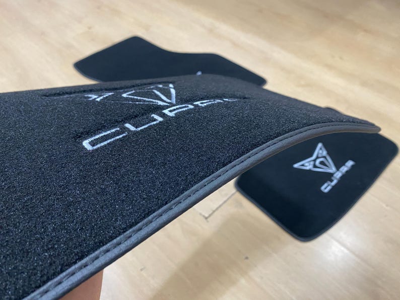 May include: Black car floor mats with the white "CUPRA" logo embroidered on the surface. The mats have a dark gray trim and are designed to protect the vehicle's interior from dirt and wear.