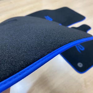 May include: Black car floor mats with a blue trim. The mats have a textured surface and a blue logo. The mats are designed to protect the car's interior from dirt and wear.