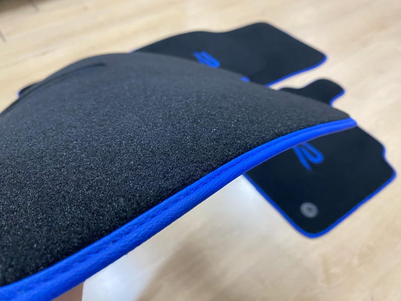 May include: Black car floor mats with a blue trim. The mats have a textured surface and a blue logo. The mats are designed to protect the car's interior from dirt and wear.