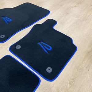 May include: Set of four black car floor mats with blue trim and a blue embroidered design. The mats have a custom shape and include mounting hardware. The mats are made of a durable material.