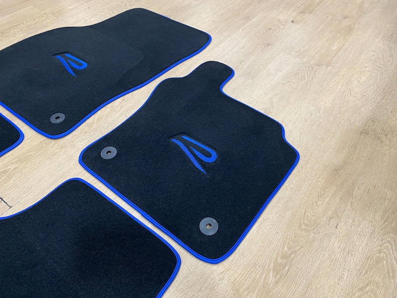 May include: Set of four black car floor mats with blue trim and a blue embroidered design. The mats have a custom shape and include mounting hardware. The mats are made of a durable material.