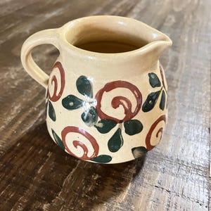 May include: A small, cream-coloured ceramic jug with a handle and spout. The jug is decorated with red spiral and green leaf designs. The jug is likely used for serving cream or other liquids.